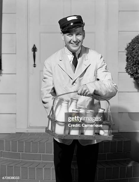 1930s SMILING MILKMAN LOOKING AT CAMERA DELIVERING MILK IN GLASS BOTTLES TO FRONT DOOR