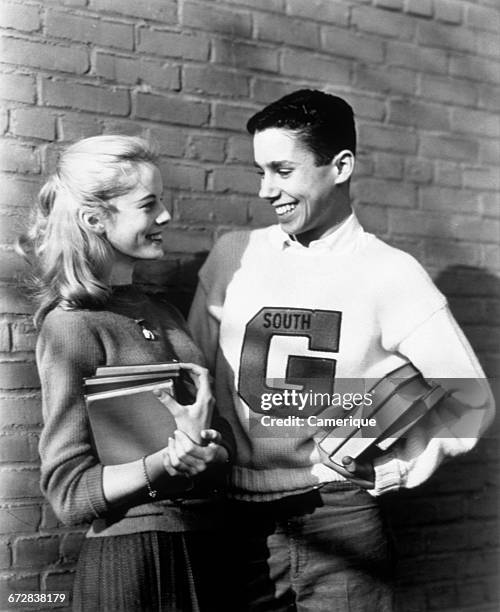 1950s TEENAGE COUPLE HOLDING BOOKS AND SMILING LEANING AGAINST WALL BOY WEARS VARSITY LETTER SWEATER
