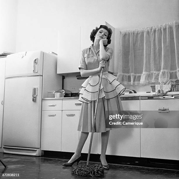 1950s DAYDREAMING BORED WOMAN HOUSEWIFE WEARING APRON LEANING ON MOP WHILE CLEANING KITCHEN FLOOR