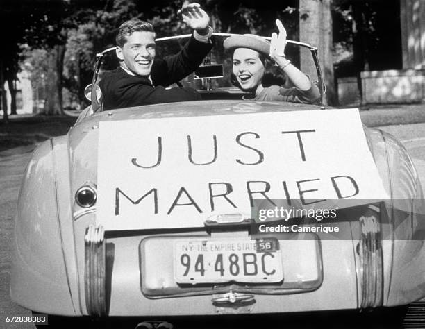 1950s NEWLYWED YOUNG COUPLE MAN WOMAN IN CONVERTIBLE SPORTS CAR WITH JUST MARRIED SIGN WAVING LOOKING AT CAMERA