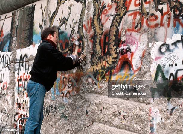 1980s TEENAGE BOY CHIPPING AWAY AT THE GRAFFITI SPRAY PAINTED BERLIN WALL WITH HAMMER AND CHISEL NOVEMBER 1989