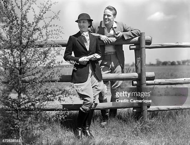 1930s COUPLE WEARING RIDING CLOTHES JODHPURS STANDING TOGETHER BY RAIL FENCE MAN SMOKING PIPE