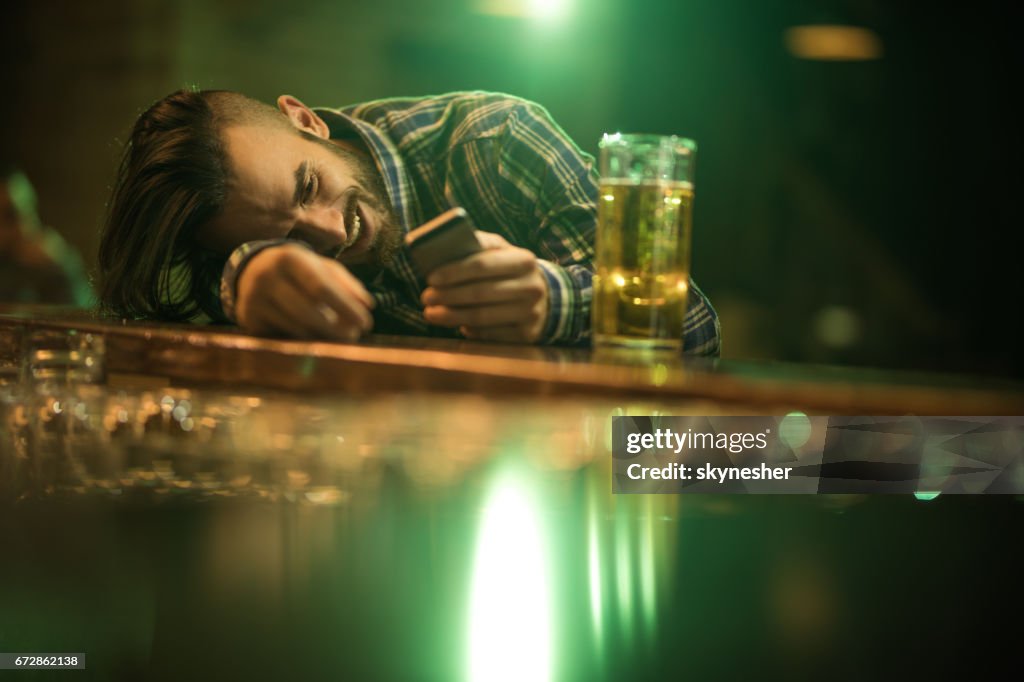 Young wasted man text messaging on cell phone while leaning on a bar counter.