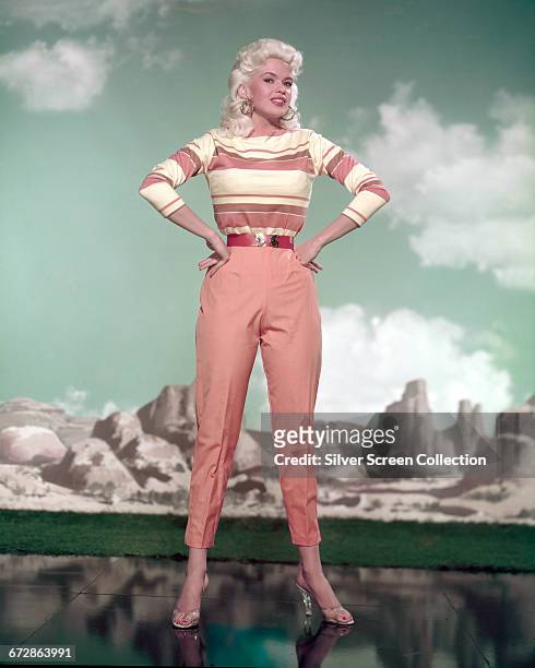 American actress Jayne Mansfield poses against a painted desert backdrop in peach-coloured trousers and a striped top, circa 1960.