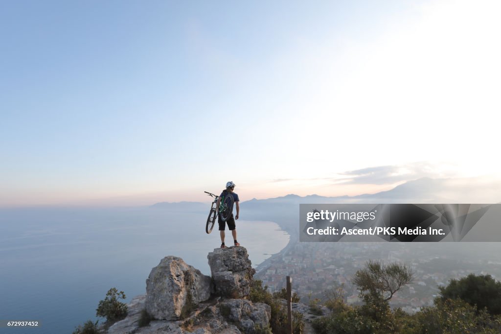 Young man carries mountain bike to summit rocks, sea below