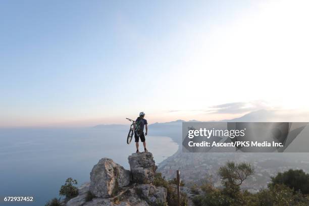 young man carries mountain bike to summit rocks, sea below - extreme sports bike stock pictures, royalty-free photos & images