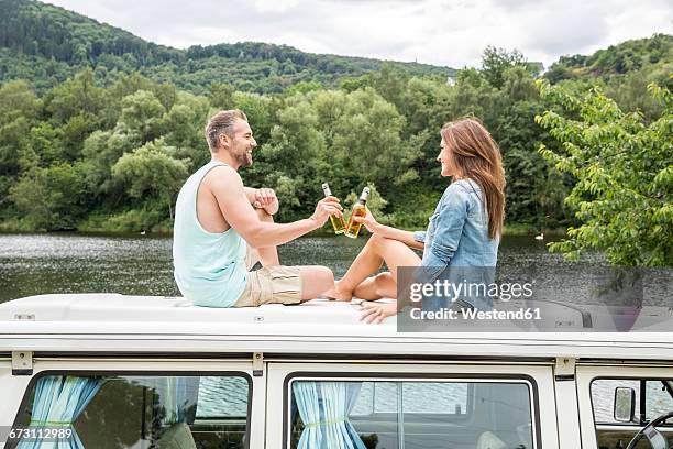 couple clinking beer bottles on roof of a van at lakeside - car roof stock pictures, royalty-free photos & images