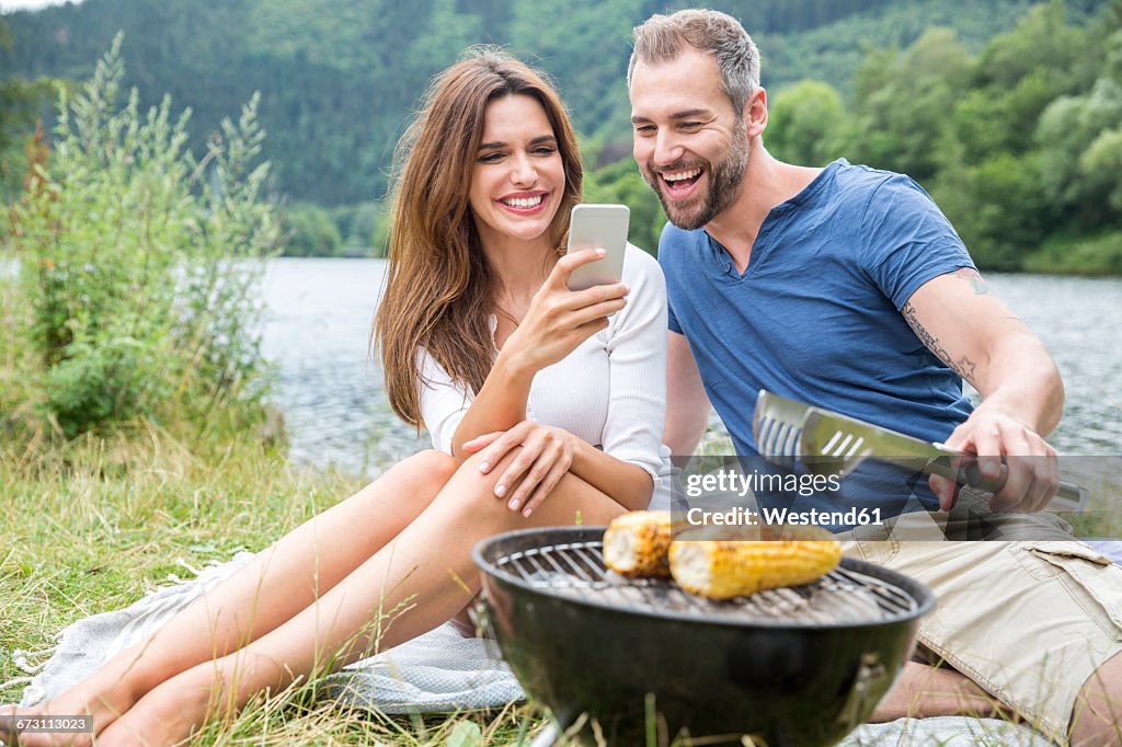 Happy couple with cell phone having a barbecue at lakeside