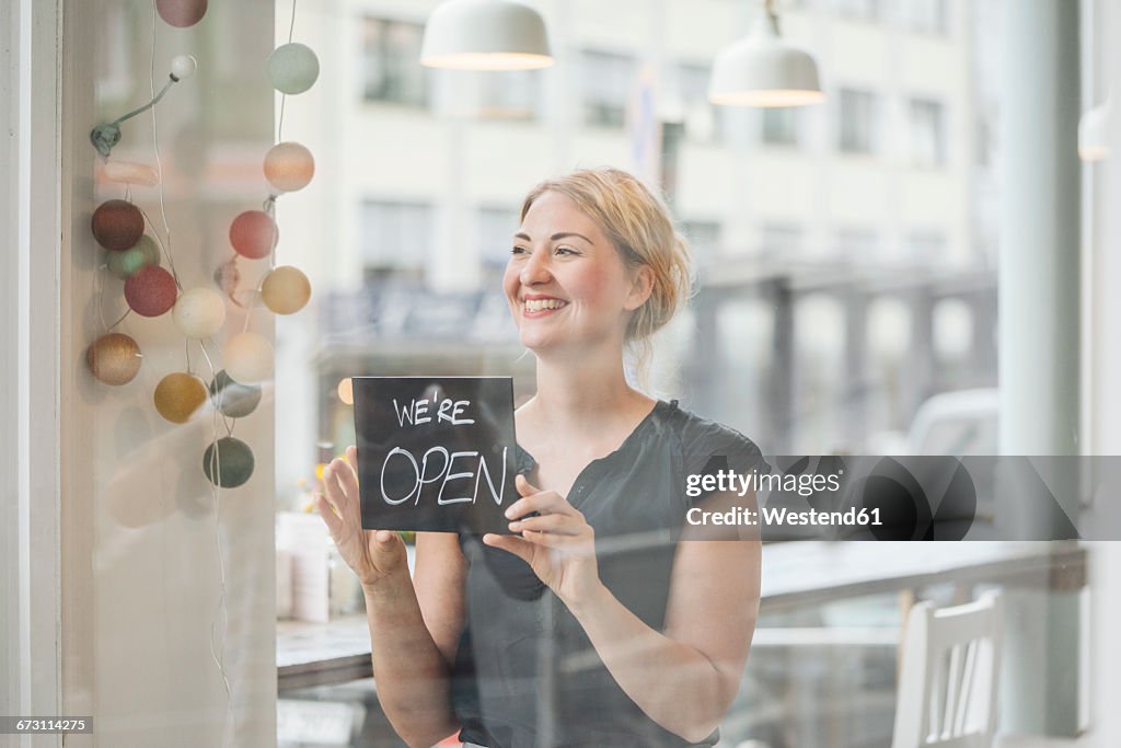 Smiling woman in a cafe attaching open sign to glass pane