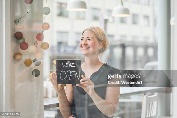 smiling woman in a cafe attaching open sign to glass pane - founder stock pictures, royalty-free photos & images