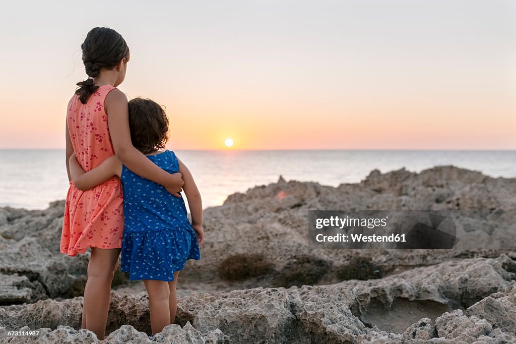 Back view of two little sisters standing arm in arm at rocky coast watching sunset