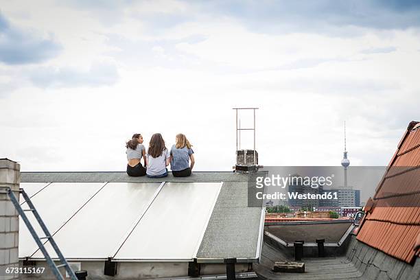 germany, berlin, back view of three friends sitting side by side on roof top - berlin fernsehturm stock-fotos und bilder