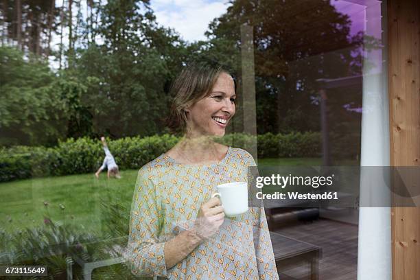 smiling woman holding cup looking out of window - window with view on garden stockfoto's en -beelden