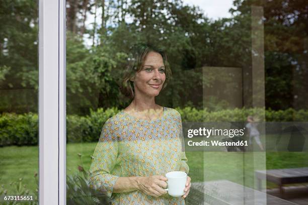 smiling woman holding cup looking out of window - window view stock pictures, royalty-free photos & images