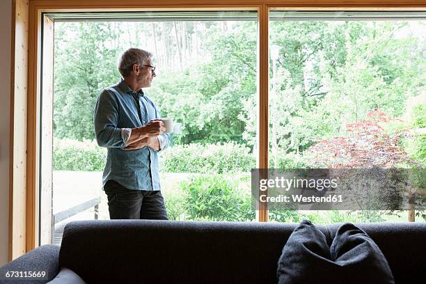 mature man holding cup looking out of window - man looking out of window stock-fotos und bilder