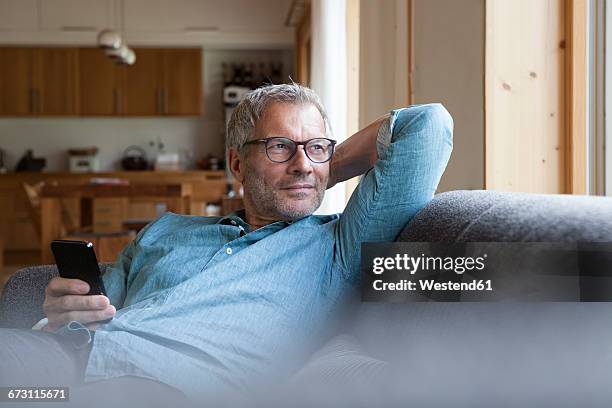 mature man holding cell phone sitting on couch - man middelbare leeftijd woonkamer stockfoto's en -beelden