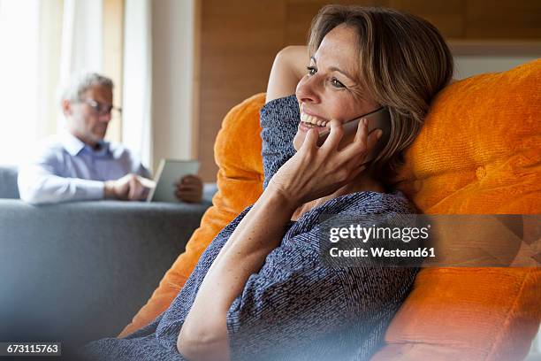 smiling woman at home on cell phone with husband in background - mature women talking stock pictures, royalty-free photos & images