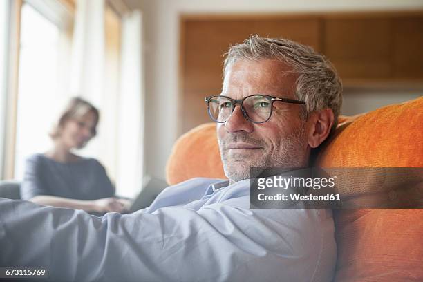 man relaxing in armchair with wife in background - ehemann stock-fotos und bilder
