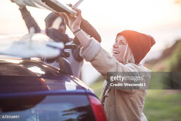 happy couple putting surfboard on car roof at sunset - car roof stock pictures, royalty-free photos & images