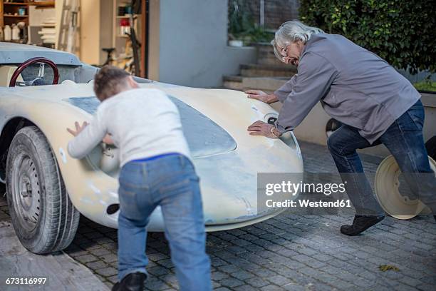 senior man and boy pushing old car back in garage - old car garage stock pictures, royalty-free photos & images