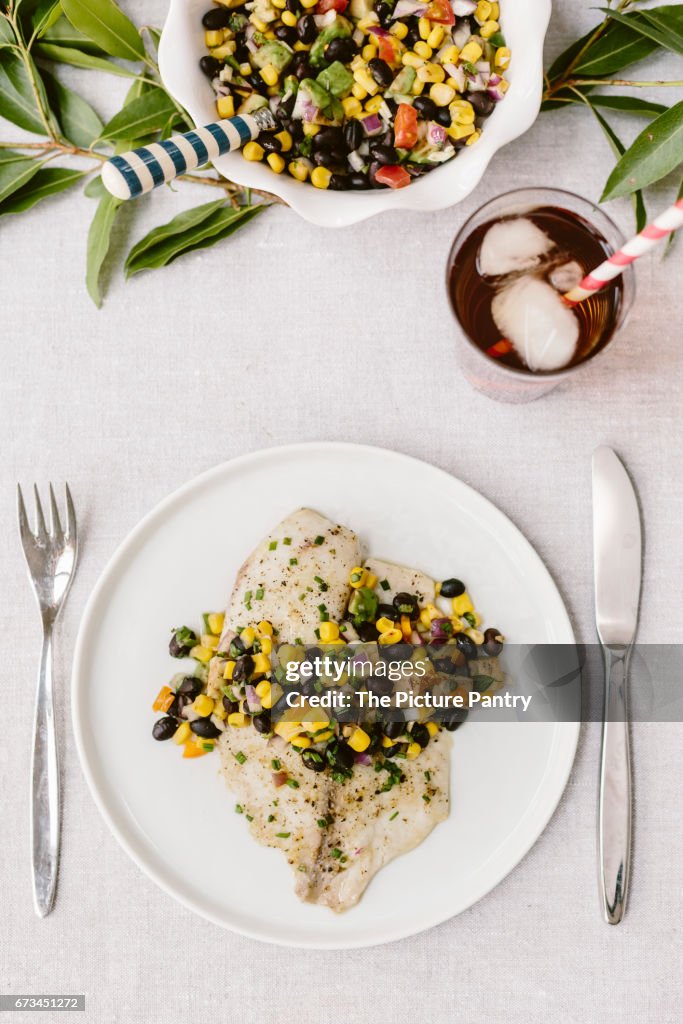 Two plates of Freshly baked tilapia topped off with black bean salsa are photographed on a summery tablesetting.
