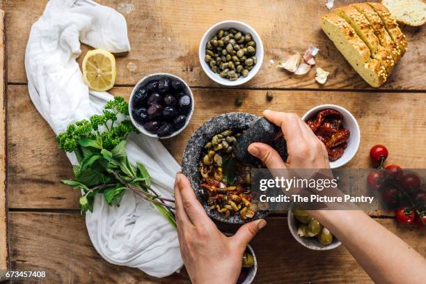 a woman mashing olives in a marble mortar to make olive tapenade is photographed from top view.  three types of olives, capers, sun-dried tomatoes herbs, corn bread slices accompany it. - olivenpasten stock-fotos und bilder