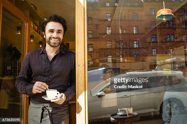 portrait of happy barista holding coffee cup while standing at doorway - barista coffee restaurant stockfoto's en -beelden