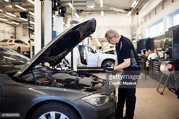 senior mechanic holding mobile phone examining car at shop - vehicle hood stock pictures, royalty-free photos & images