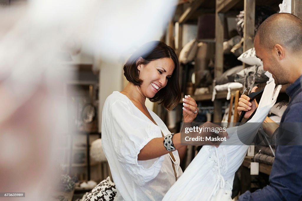 Smiling female customer looking at white dress held by owner in store