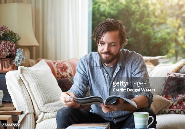 man reading magazine on sofa at home. - uomini di età media foto e immagini stock