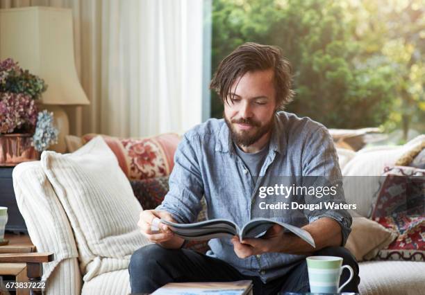 man reading magazine on sofa at home. - mid adult men stock pictures, royalty-free photos & images