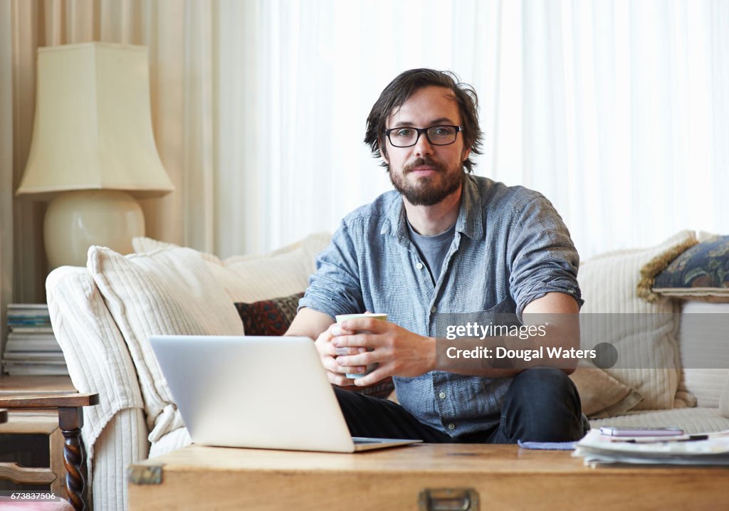 Portrait of man sitting on sofa with laptop and hot drink.