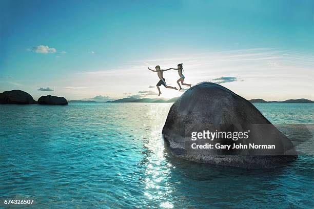 two kids holding hands jumping off rock into sea - sunny side fotografías e imágenes de stock