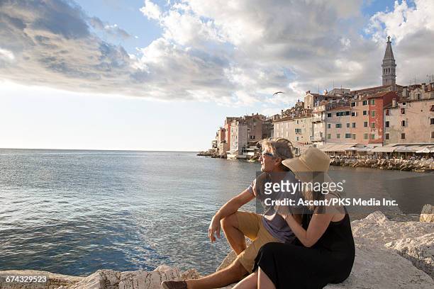 couple rest on rocks above sea, town behind - woman look straight black shirt stock pictures, royalty-free photos & images