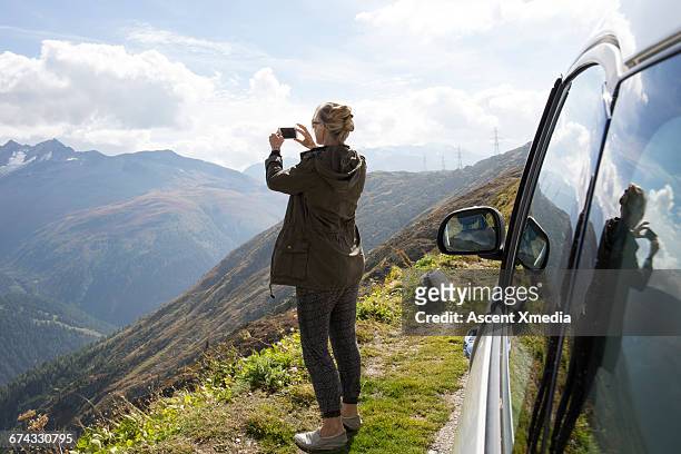 woman takes smart phone pic beside car, mtns - rear view photos stockfoto's en -beelden