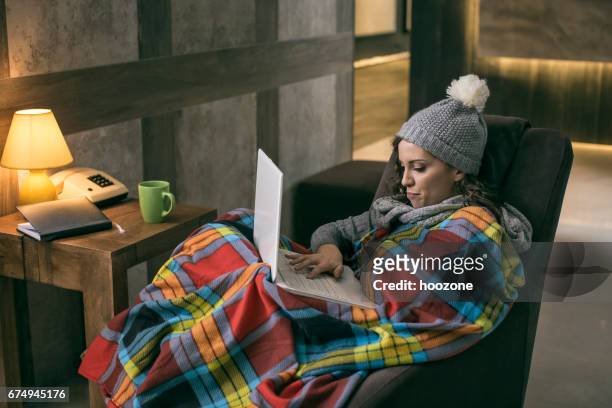 femmes portant hiver cap travail sur ordinateur portable, enveloppé dans une couverture à la maison avec pas de chauffage - froid photos et images de collection
