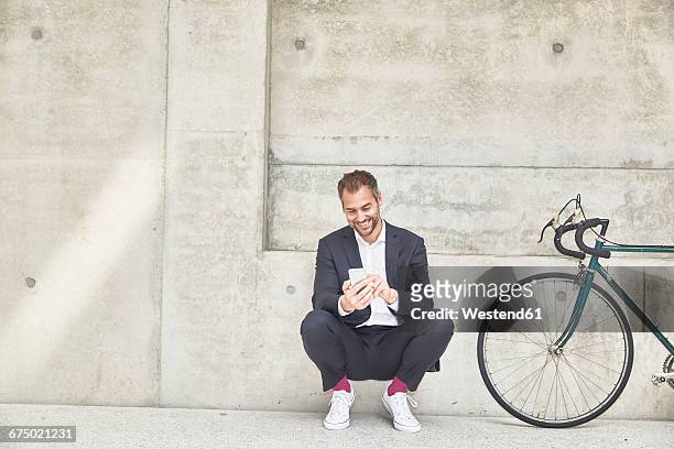 smiling businesssman with bicycle at concrete wall looking at cell phone - hombre agachado fotografías e imágenes de stock