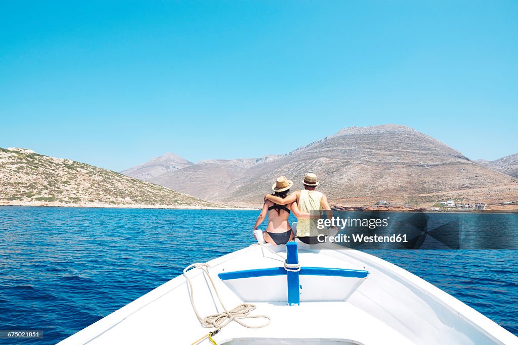 Greece, back view of couple sitting arm in arm on bow of a boat looking at Amorgos Island