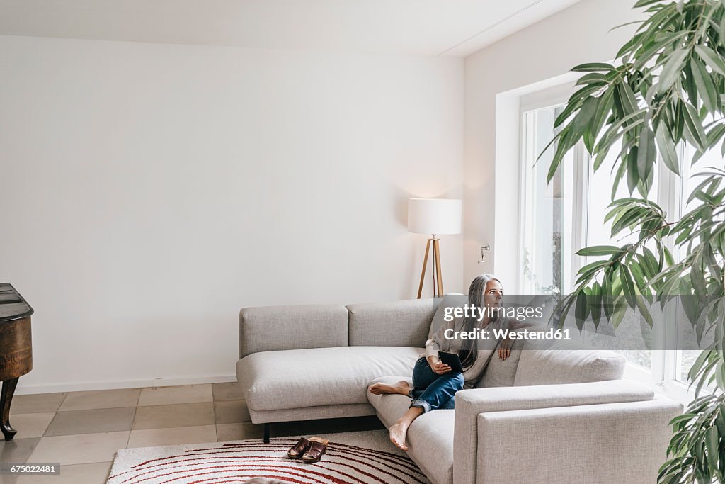 Pensive woman sitting with mini tablet on the couch looking through window