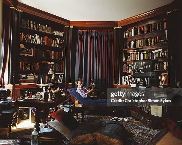 English guitarist Keith Richards of the Rolling Stones playing a guitar on a chaise longue in his library, circa 1995.