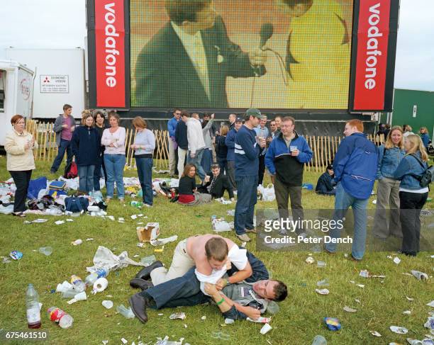 Two young men fool about on the grass beneath a screen showing the action on Derby Day at Epsom Downs Racecourse. June 2001.