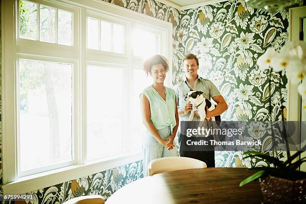 smiling couple holding dog in dining room of home - black white floral wallpaper stock-fotos und bilder