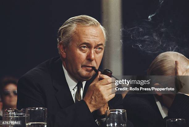 British politician and leader of the Labour Party, Harold Wilson smokes a pipe as he listens to a speech on the platform at the Labour Party annual...