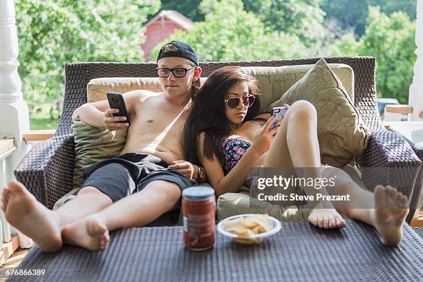teenage couple wearing bikini and swim shorts reclining on patio sofa reading smartphones - teen boy shorts stock-fotos und bilder