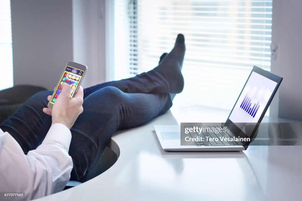 Lazy office worker playing mobile game with smartphone during work hours. Avoiding his job and being lazy with feet   and socks on table. Useless and relaxing man doing nothing and forget his job.