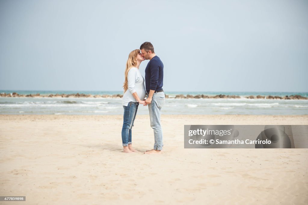 Pregnant couple kissing on the beach. Hand in hand. Casual clothes.