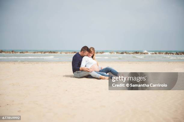 pregnant couple sitting on the beach. embrace. casual clothes. - relazione di coppia stockfoto's en -beelden