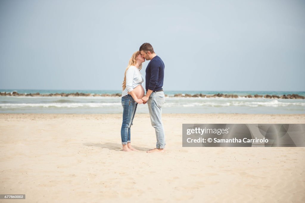 Pregnant couple kissing on the beach. Hand in hand. Casual clothes.