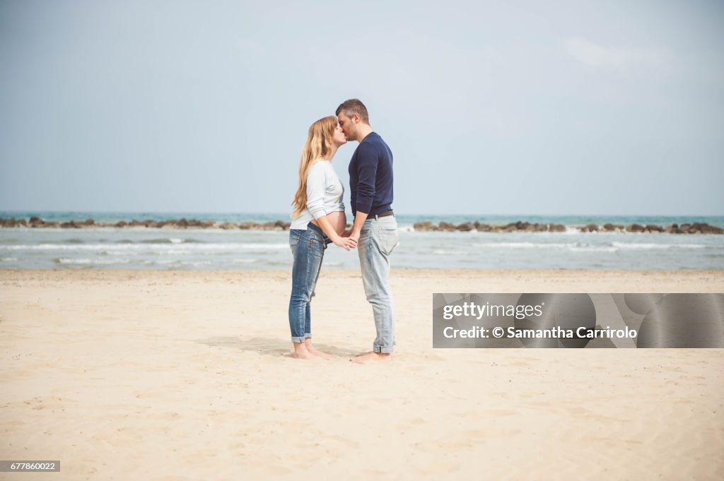Pregnant couple kissing on the beach. Hand in hand. Casual clothes.