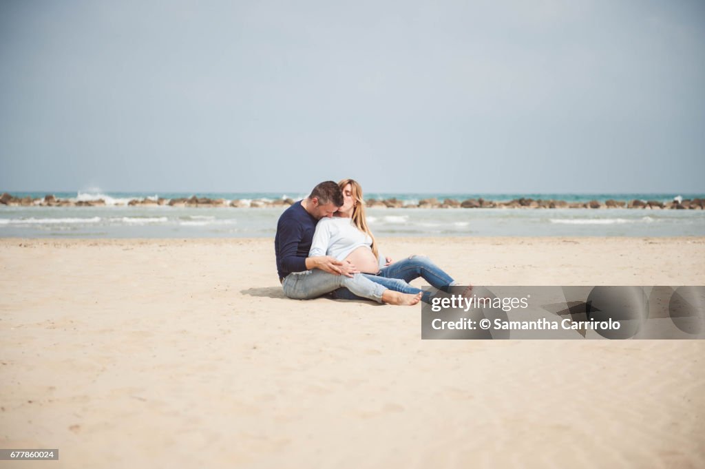 Pregnant couple sitting on the beach. Embrace. Casual clothes.